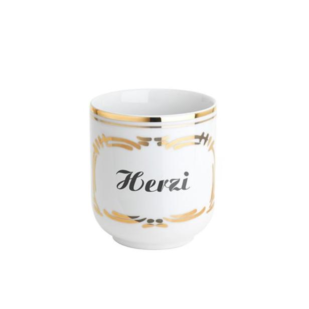 Eine weiße Tasse mit goldenen Verzierungen und dem Schriftzug „Herzi“.