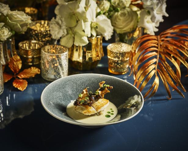 Ein Teller mit Fisch, garniert mit Pilzen und Kräutern, auf einem festlich gedeckten Tisch.