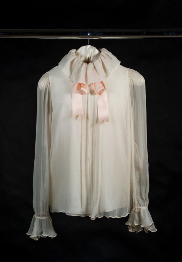 Eine weiße Bluse mit Rüschenkragen und rosa Schleife hängt an einem Kleiderbügel.