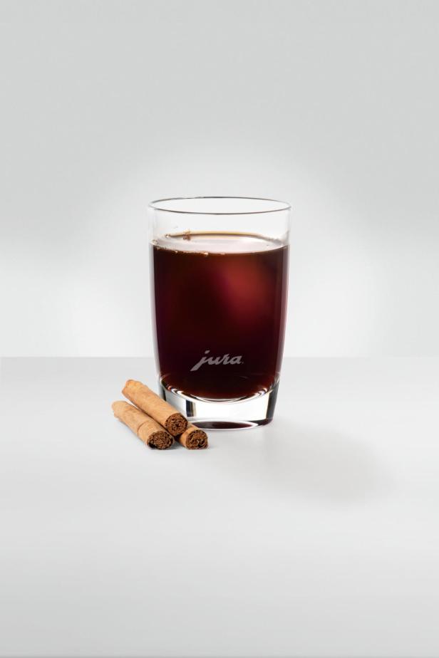 Ein Glas mit dunklem Kaffee und drei Zimtstangen auf einem weißen Tisch.