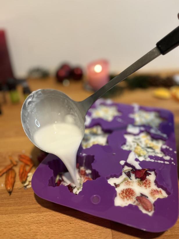 Mit einer Schöpfkelle wird weiße Flüssigkeit in eine lila Silikonform gegossen, die mit Blüten gefüllt ist.