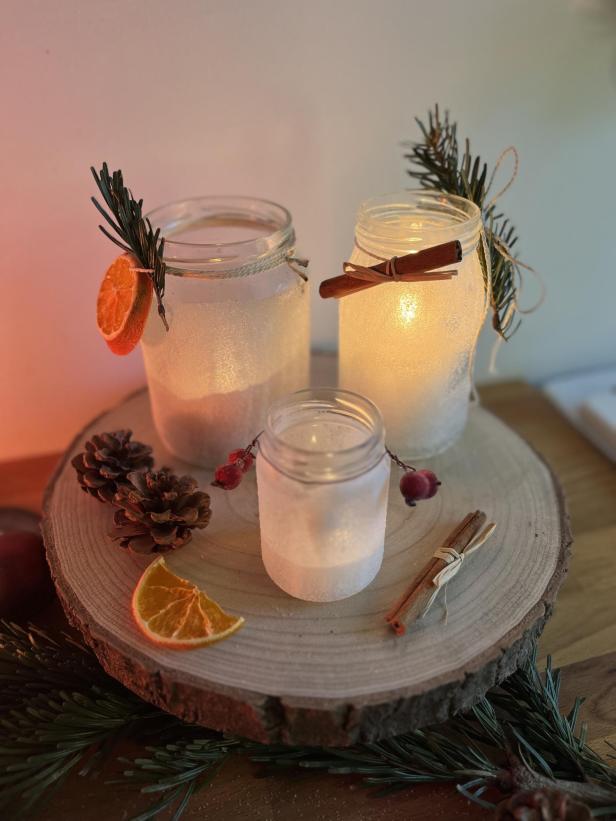 Drei dekorative Windlichter in Gläsern mit Zimtstangen und Orangenscheiben auf einer Holzscheibe.