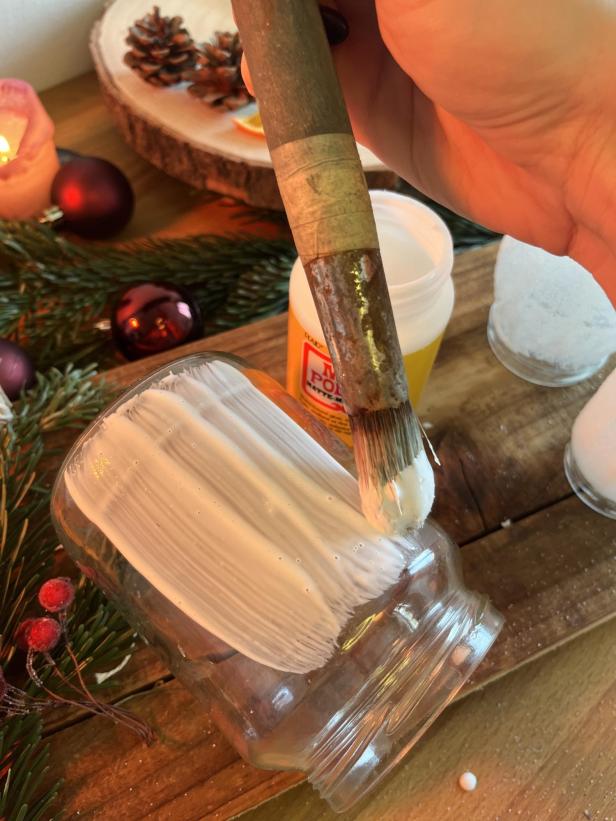 Ein Glas wird mit einem Pinsel und weißer Farbe bemalt, um eine winterliche Dekoration zu gestalten.
