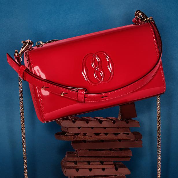 Eine rote Lackhandtasche liegt auf einem Stapel dunkler Schokolade vor blauem Hintergrund.