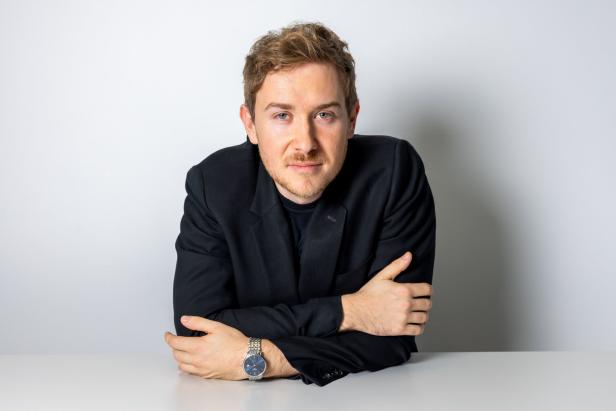 Ein Mann mit braunen Haaren und einem schwarzen Sakko sitzt an einem Tisch.