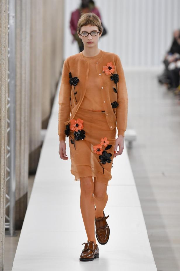 Ein Model präsentiert ein orangefarbenes Kleid und eine Strickjacke mit Blumenapplikationen von Miu Miu.
