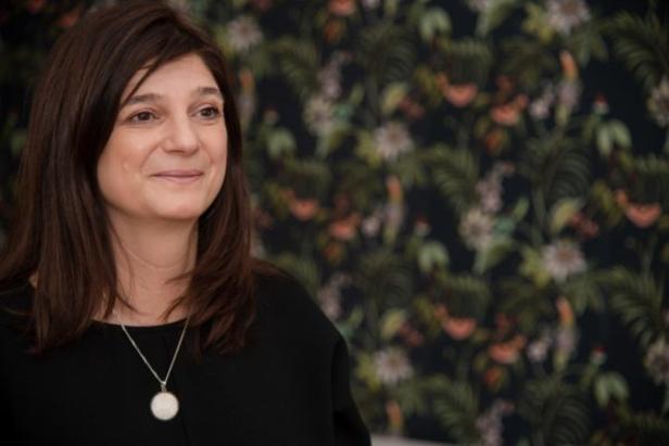 Eine Frau mit braunen Haaren und einer Halskette vor einem floralen Hintergrund.
