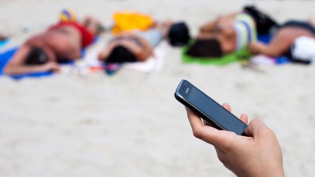 Eine Hand hält ein Smartphone am Strand, im Hintergrund liegen mehrere Menschen in der Sonne.
