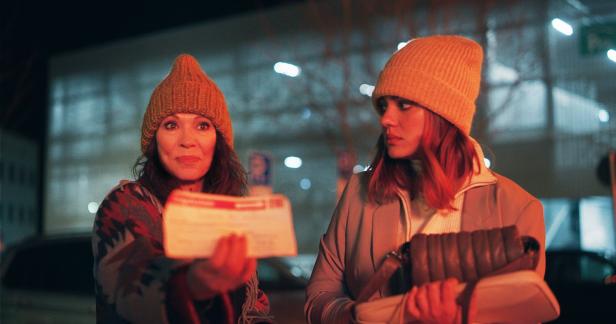 Zwei Frauen mit Mützen stehen nachts auf der Straße, eine hält einen Brief in der Hand.