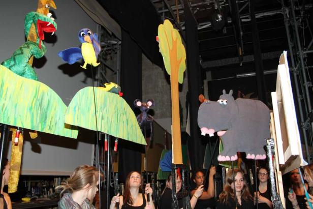 Mehrere Personen bedienen Stabpuppen, darunter eine Schlange, ein Vogel und ein Nilpferd.