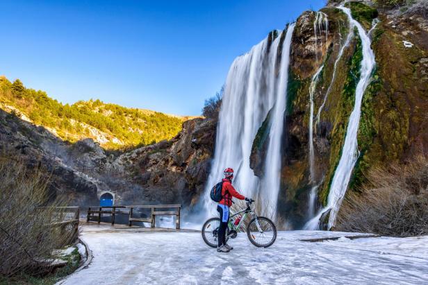 Eine Radfahrerin steht vor einem hohen Wasserfall in einer bergigen Landschaft.