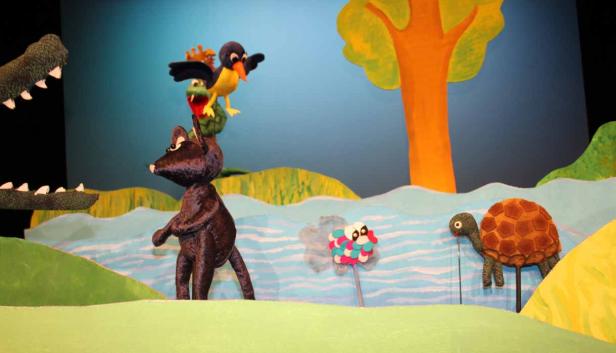 Eine Puppenspielszene mit einem Bären, einem Krokodil, Vögeln und einer Schildkröte vor einem gemalten Hintergrund.