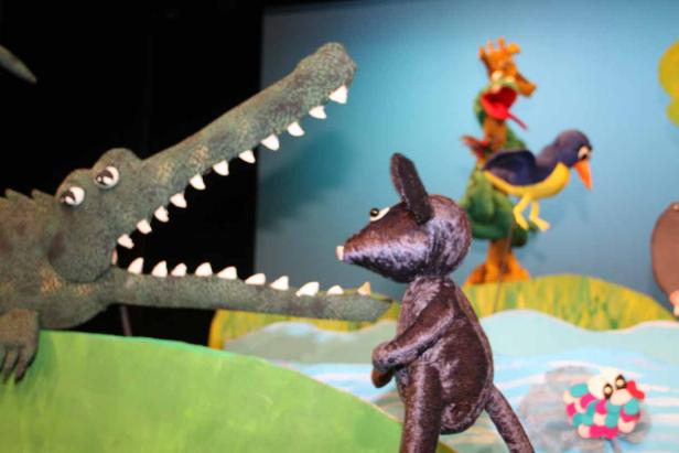 Ein Krokodil mit offenem Maul steht vor einer kleinen Maus in einer Puppentheater-Szene.