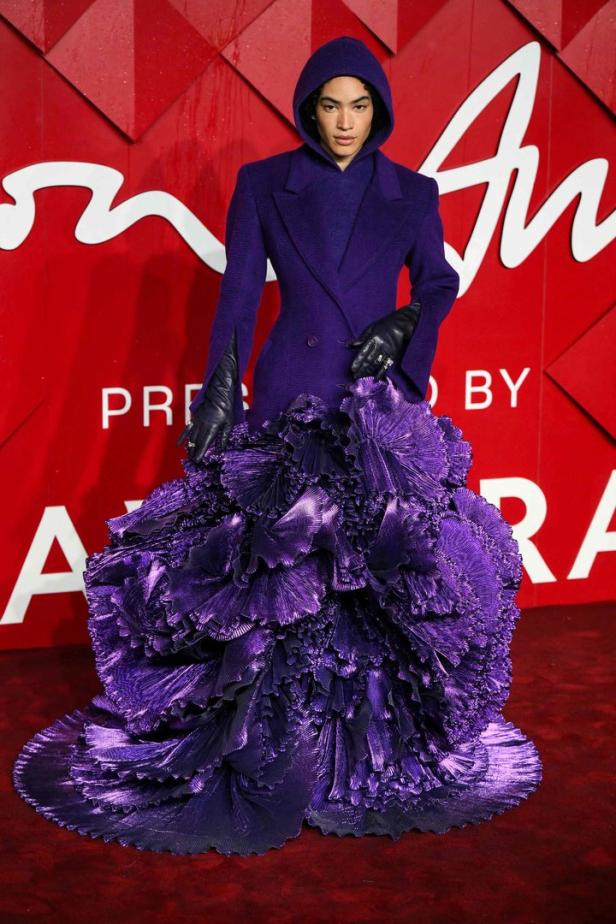 Eine Person in einem auffälligen, violetten Kleid mit Kapuze posiert vor einem roten Hintergrund.
