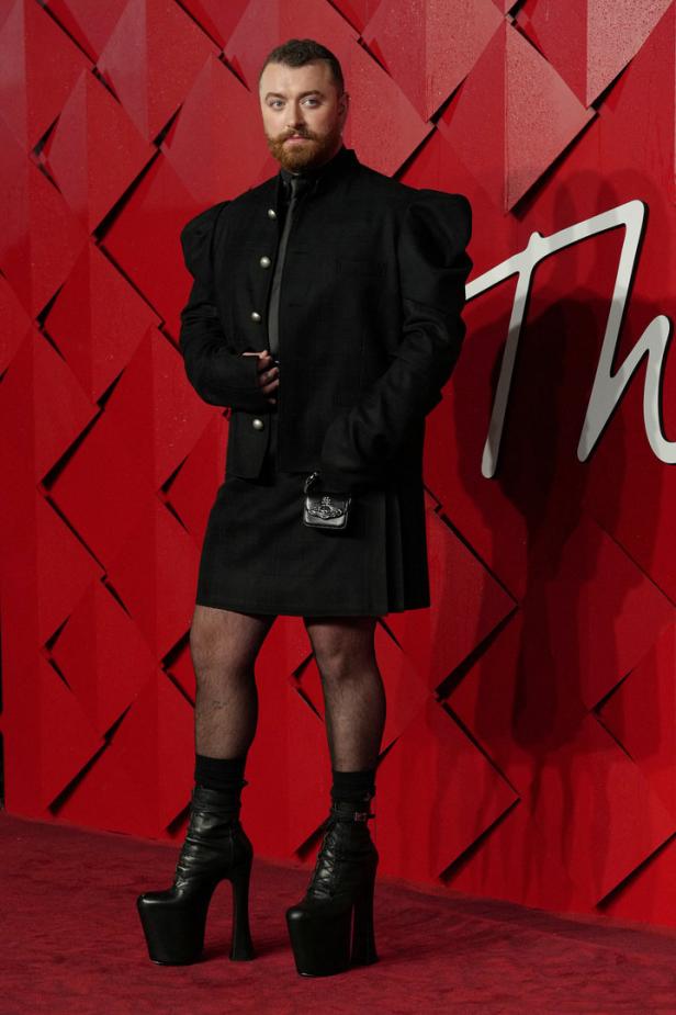 Sam Smith trägt einen Rock, eine Jacke mit Puffärmeln und Plateaustiefel vor einem roten Hintergrund.