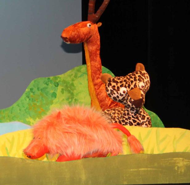 Drei Stofftiere, eine Giraffe, ein Leopard und ein Wildschwein, stehen auf einer Bühne.
