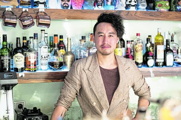 Kan Zuo, bekannt aus „The Sign“, steht vor einer Bar mit Spirituosenregalen.