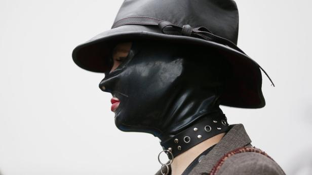 Eine Person mit rotem Lippenstift trägt eine schwarze Ledermaske und einen Hut.