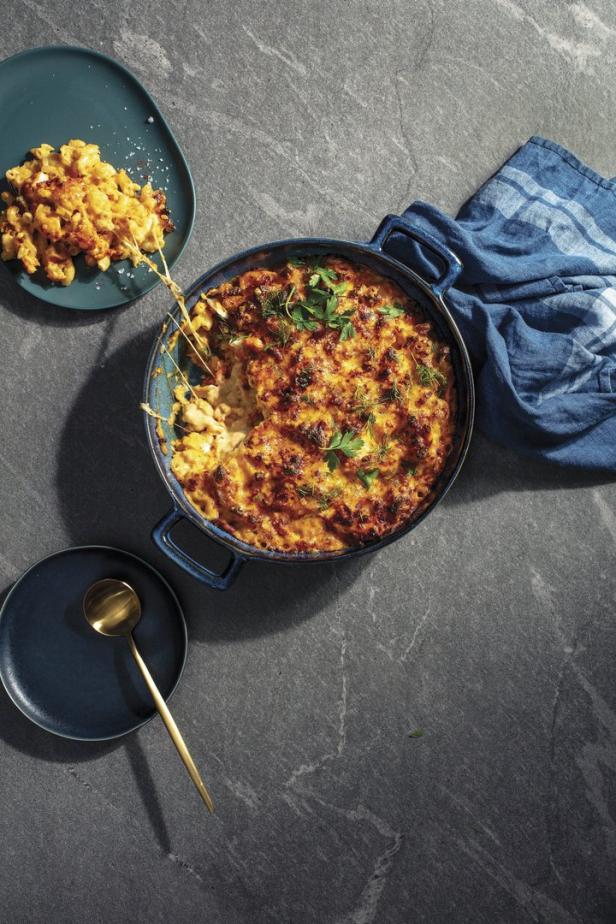 Ein Auflauf mit Mac and Cheese wird mit einem goldenen Löffel und einem blauen Geschirrtuch präsentiert.