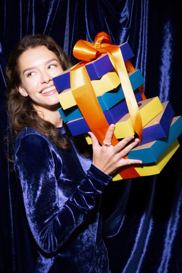 Eine lächelnde Frau im blauen Samtkleid hält einen Stapel bunter Geschenke mit orangefarbenen Schleifen.