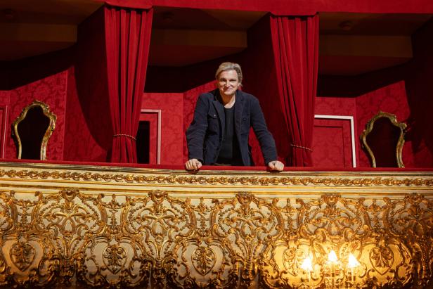 Ein Mann steht in einer Theaterloge mit roten Vorhängen.