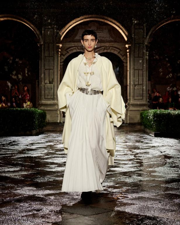 Ein Model präsentiert ein weißes Kleid und einen cremefarbenen Mantel auf einem nassen Laufsteg.