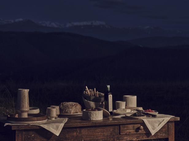 Ein gedeckter Tisch mit Brot, Käse und Olivenöl vor einer Bergkulisse bei Nacht.