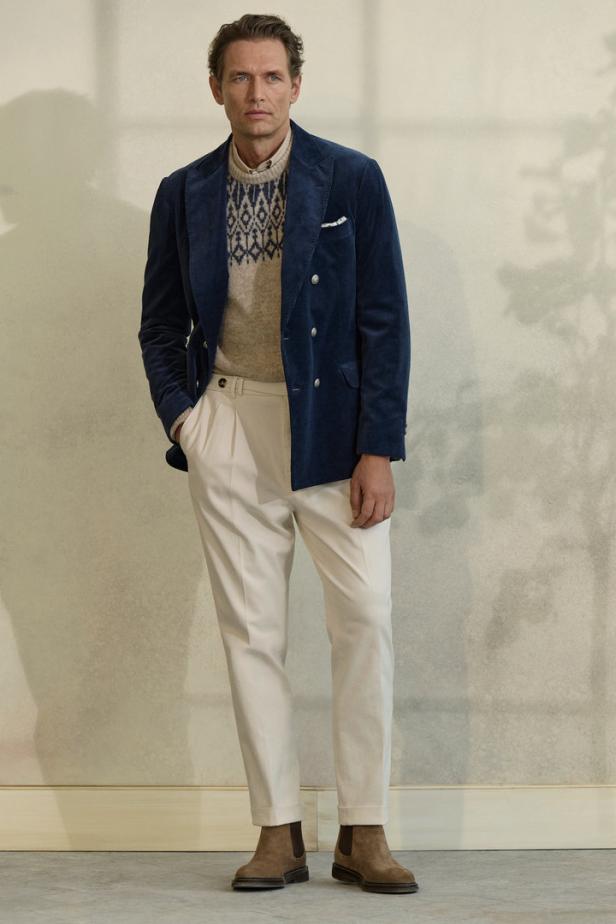 Ein Mann trägt ein blaues Samtsakko, einen gemusterten Pullover, weiße Hosen und braune Stiefel.