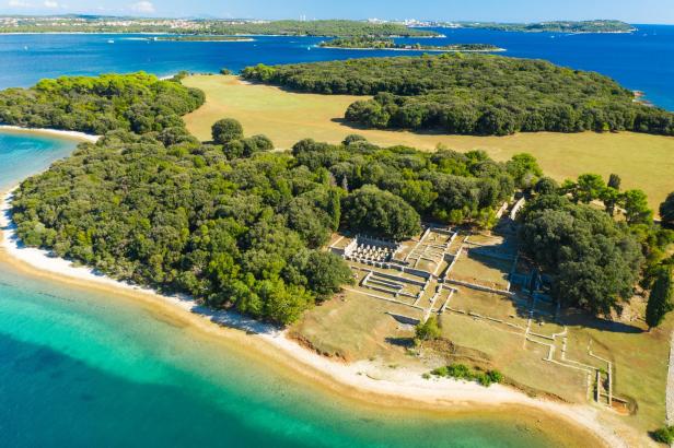 Luftaufnahme der Ruinen einer römischen Villa auf einer bewaldeten Insel mit türkisfarbenem Wasser.