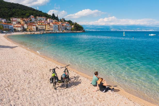 Ein Paar sitzt am Strand neben ihren Fahrrädern und blickt auf das Meer.
