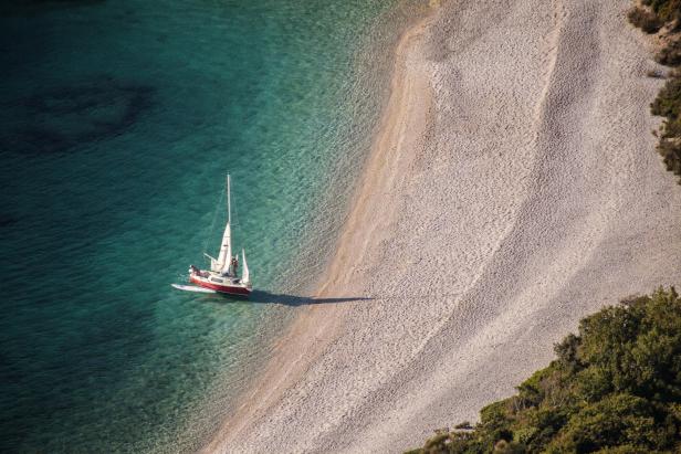 Ein rotes Segelboot ankert vor einem Kiesstrand mit türkisfarbenem Wasser.