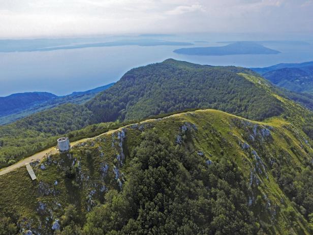 Ein bewaldeter Bergrücken mit einem kleinen Turm und Blick auf einen See.