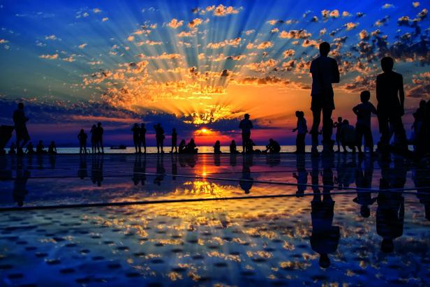 Menschen beobachten den Sonnenuntergang am Meer, ihre Silhouetten spiegeln sich auf dem Boden.