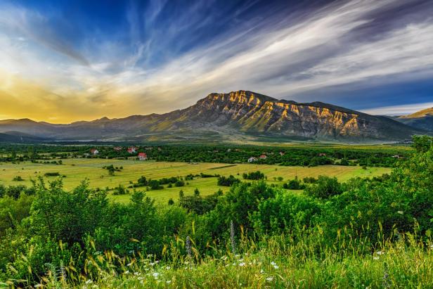 Eine grüne Landschaft mit einem Dorf und einem Berg unter einem bewölkten Himmel.
