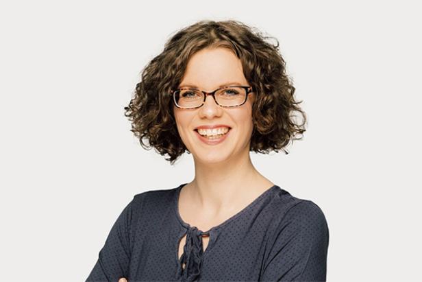 Eine lächelnde Frau mit Brille und lockigem Haar vor einem hellen Hintergrund.