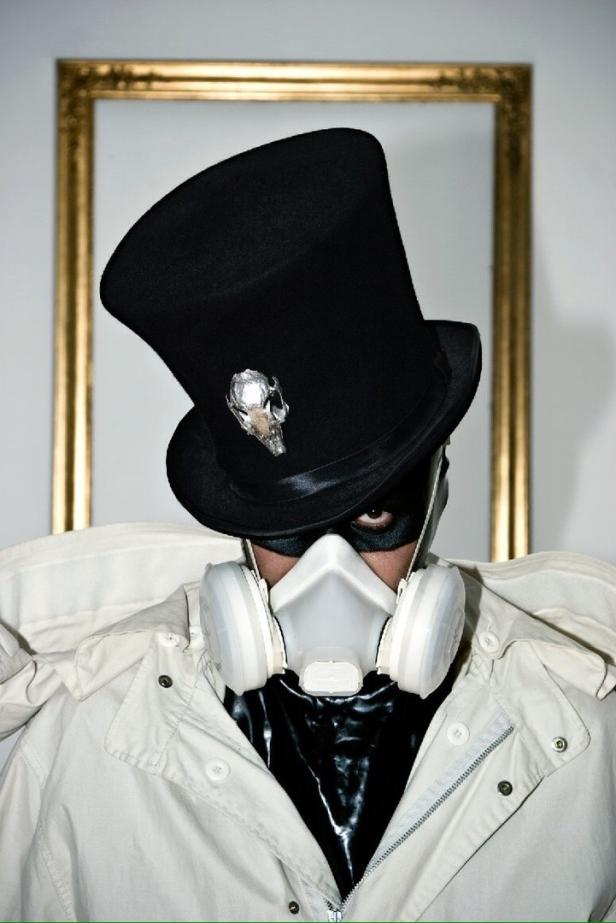Eine Person mit Zylinder, Maske und Atemschutzmaske vor einem goldenen Rahmen.