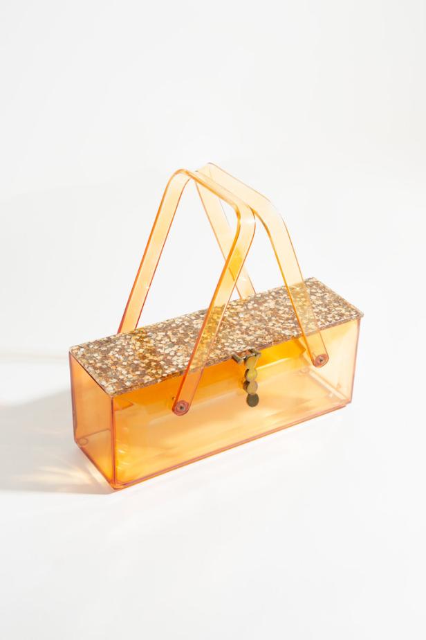 Eine orangefarbene Handtasche aus Kunststoff mit goldenem Glitzerdeckel steht vor einem weißen Hintergrund.
