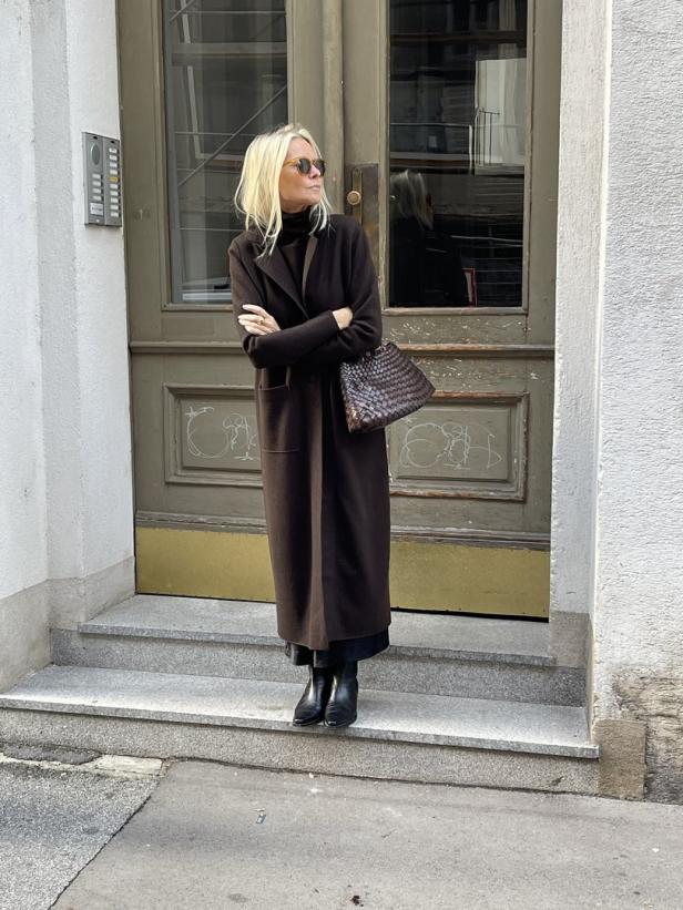 Eine blonde Frau mit Sonnenbrille und braunem Mantel steht vor einer Tür.