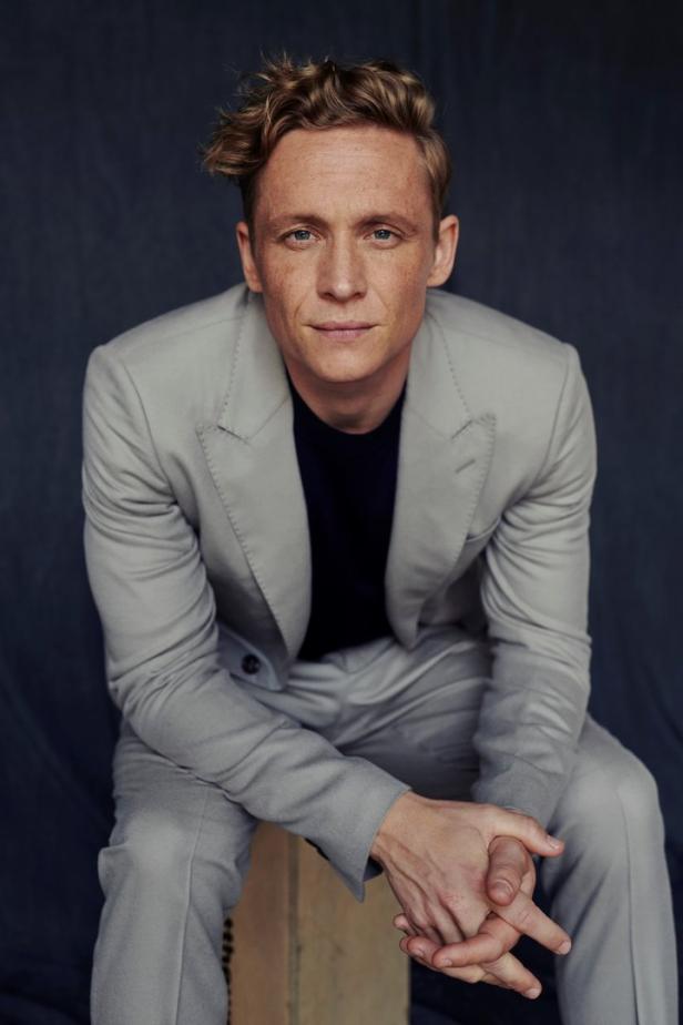 Ein Mann mit blonden Haaren und einem grauen Anzug sitzt vor einem dunklen Hintergrund.