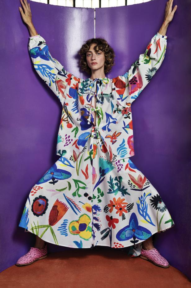 Eine Frau in einem farbenfrohen, floral gemusterten Outfit posiert vor einem lila Hintergrund.