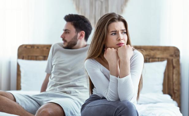 Mann und Frau sitzen getrennt auf einem Bett, die Frau wirkt besorgt.
