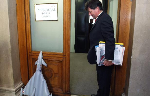 Ein Mann betritt einen Raum mit der Aufschrift „Budgetsaal“, vor der Tür steht ein aufblasbares Flugzeug.