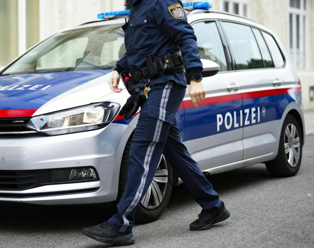 Ein Polizist in Uniform geht an einem silbernen Polizeiauto mit blauen und roten Streifen vorbei.