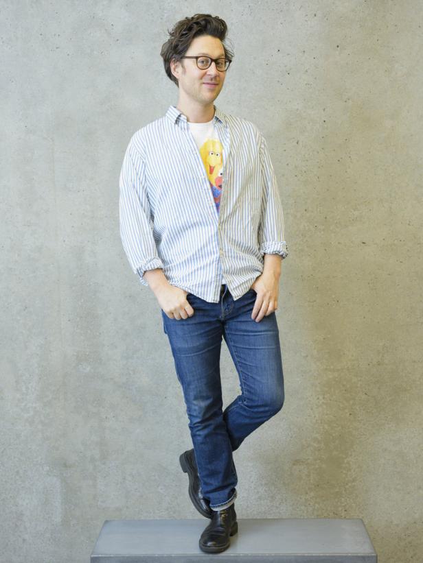 Ein Mann mit Brille, Jeans und Hemd posiert vor einer grauen Wand.