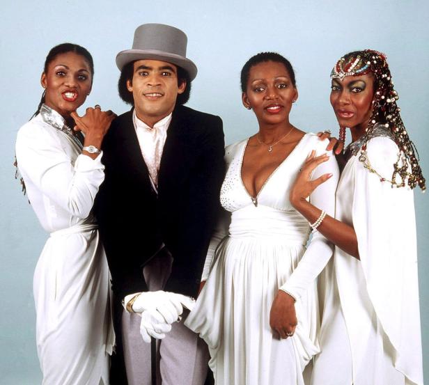 Die Musikgruppe Boney M. posiert in weißen Outfits und mit einem Zylinderhut für ein Foto.