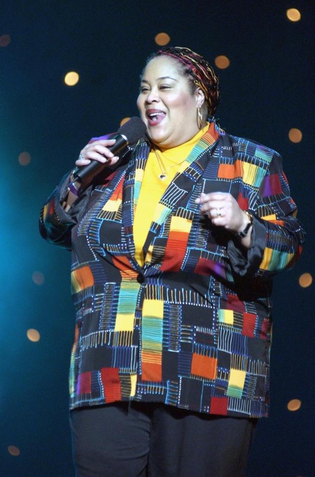 Eine Frau mit einem bunten Blazer singt in ein Mikrofon auf einer Bühne.
