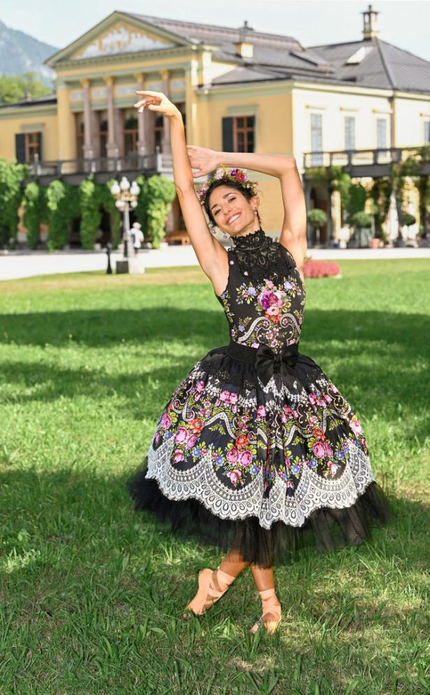 Eine Ballerina in einem farbenfrohen Kleid posiert auf einer Wiese vor einem gelben Gebäude.