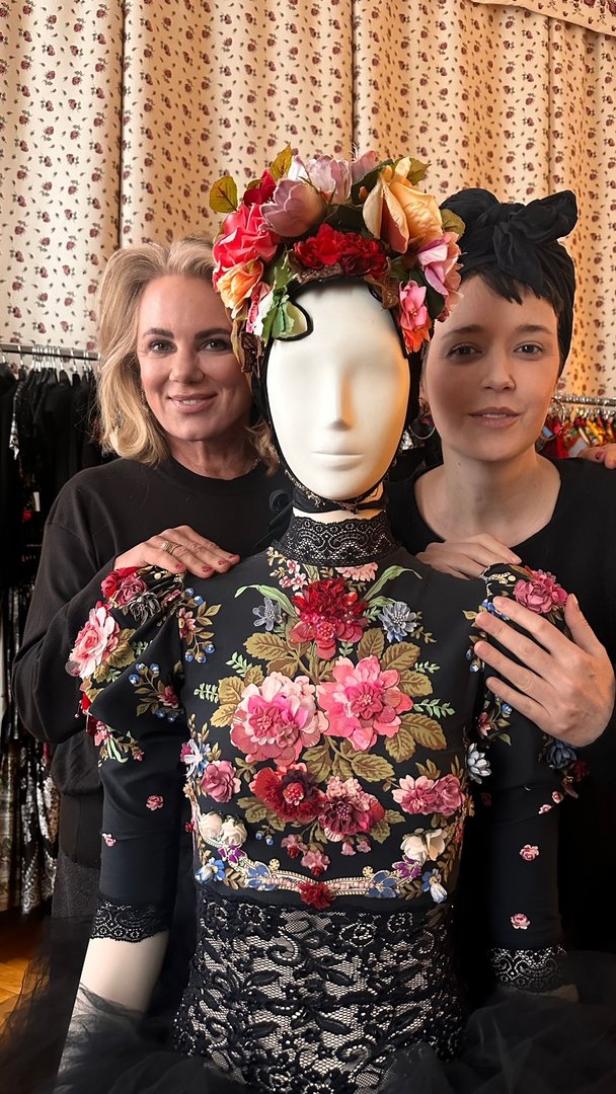 Zwei Frauen posieren neben einer Schaufensterpuppe mit einem aufwändigen Blumenkleid und Kopfschmuck.