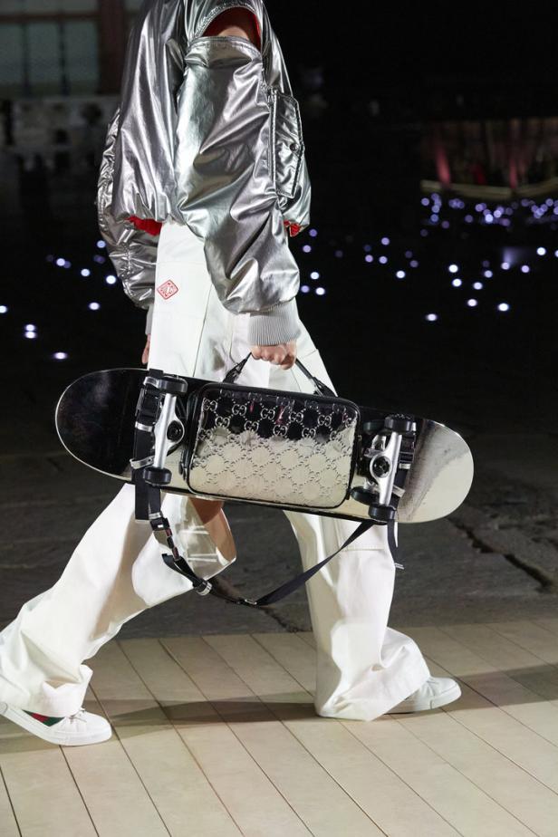 Ein Model trägt eine silberne Jacke und eine Gucci-Tasche, die an einem Skateboard befestigt ist.