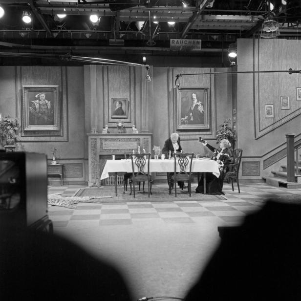 Zwei Personen sitzen an einem Tisch in einem Fernsehstudio, umgeben von Kameras und Scheinwerfern.